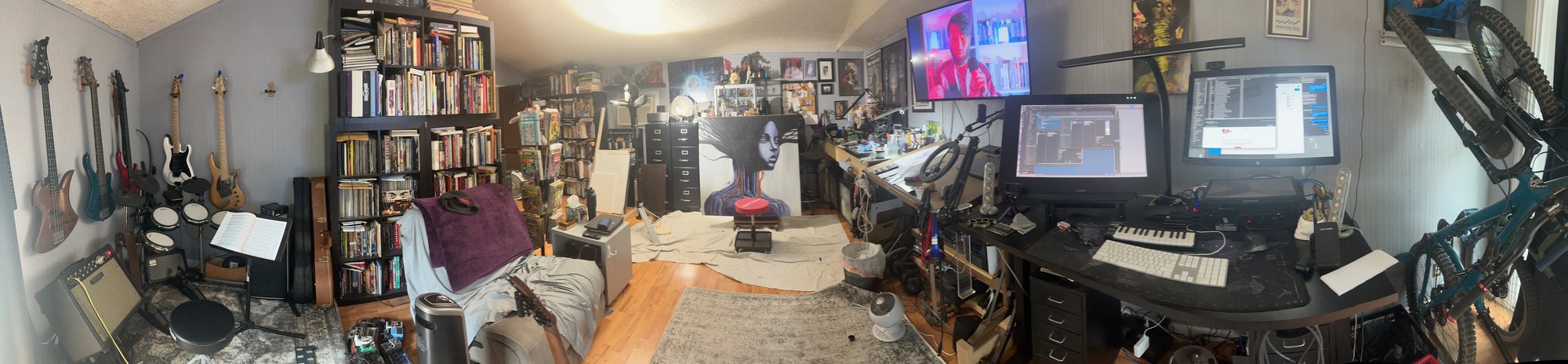 A panoramic view of Ari’s home studio: bass guitars and drums on the left, bookshelves, a large painting on an easel, and a standing desk with monitors and a keyboard on the right.